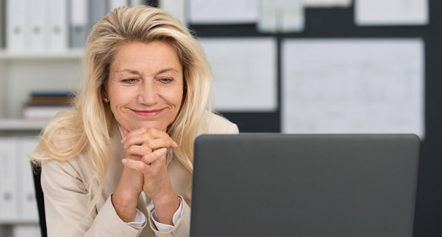 A woman in an office sitting at her laptop. She has her hands clasped beneath her chin and is smiling.