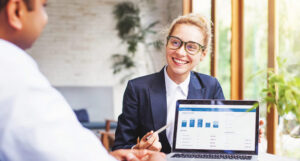 A business woman sits at a desk, presenting a proposal (on a laptop screen) to the man sitting across from her.