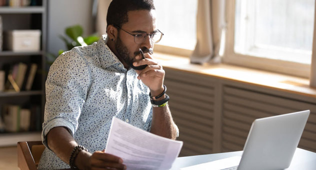 A man with some papers in his hand sitting at a desk. He is focused intently on his laptop screen.