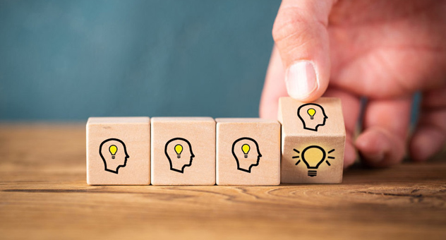 Four wooden cubes on a table. Each block shows the profile of a person's head with a small lightbulb inside. A hand turns the last block over, revealing a bigger lightbulb on the other face of the cube.