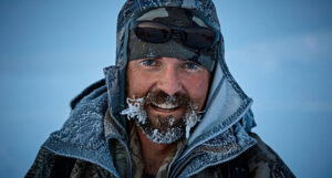 A man standing in an icy landscape wearing a winter hat and coat. He has icicles forming on his moustache and beard.