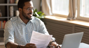 A man sitting at a desk in his home office. He's holding a report document and looking at his laptop.