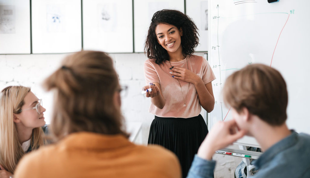 Professional medium-skinned woman at whiteboard gives presentation to colleagues. She smiles and gestures towards a person in the audience.