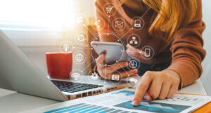 A woman at a desk pointing at a strategy document. She has her phone in hand and icons like a shopping card, camera, chat bubbles, etc radiate out from the phone.