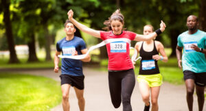 Woman in a red t-shirt and black leggings winning a race in a park. There are three runners behind her as she breaks triumphantly through the finish line.